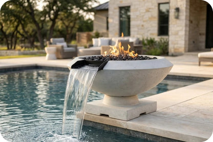 A modern concrete fire pit bowl with a waterfall feature flowing into a pool, set on a patio in front of a stone house.