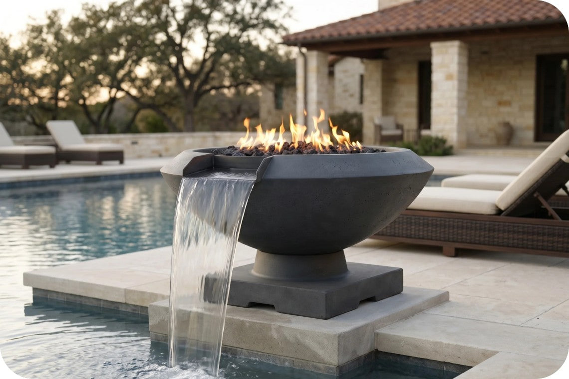 A modern outdoor fire pit with a bowl-shaped dark gray stone base, featuring flowing water from a top spout into a pool and burning flames inside, set on a stone deck beside a swimming pool at dusk.