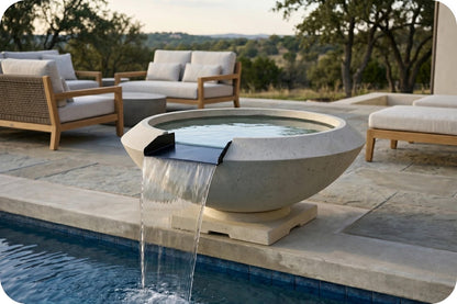 A round concrete outdoor fountain with a waterfall spout, placed beside a swimming pool on a stone patio with lounge chairs and trees in the background.