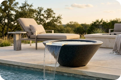 A modern outdoor poolside scene with a large dark stone bowl-shaped feature wall fountain with water cascading into the pool, set on a light stone patio with a lounge chair and trees in the background.