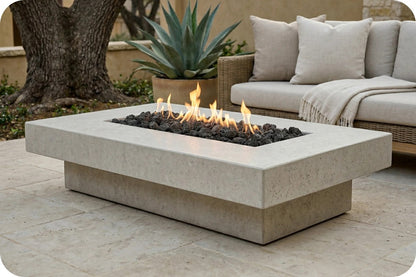 A rectangular outdoor fire pit table with a light beige stone-look body, set on a patio with black fire rocks and visible flames. A cushioned outdoor sofa, potted plant, and tree are visible in the background.