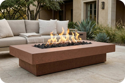 A rectangular outdoor fire pit table with a warm terracotta-brown concrete finish, filled with black river rocks and active orange flames, set on a stone patio in front of a modern outdoor sofa.