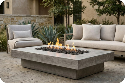 A modern rectangular concrete fire pit table with black stones and visible flames, placed outdoors between a light beige armchair and a matching sofa on a stone patio.