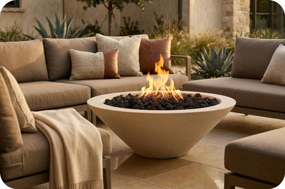 A round concrete fire pit with black river rocks and a visible flame, placed in front of a beige outdoor sectional sofa on a patio.