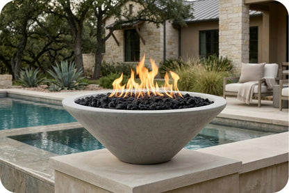A modern concrete fire pit bowl filled with black decorative stones and active orange flames, situated on a stone patio beside a swimming pool with a house and outdoor seating in the background.