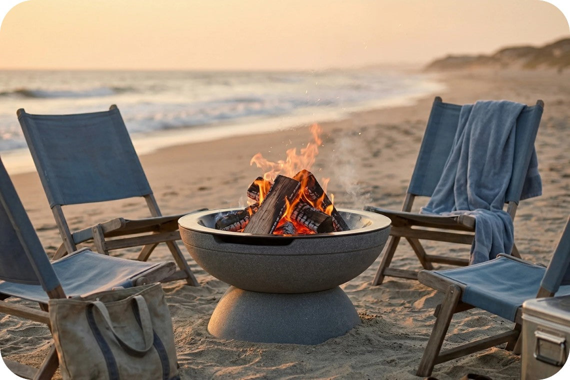 A round concrete fire pit with a burning wood fire sits on a sandy beach, surrounded by four blue lounge chairs with a blue towel draped over one chair, set against a sunset ocean background.