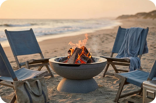 A round concrete fire pit with a burning wood fire sits on a sandy beach, surrounded by four blue lounge chairs with a blue towel draped over one chair, set against a sunset ocean background.