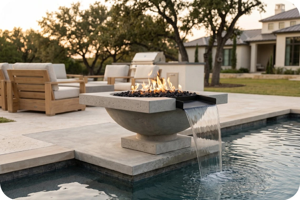 A modern outdoor fire pit with a waterfall feature, set in a pool deck next to a swimming pool. The spherical concrete fire bowl is lit with flames, with water cascading down a black ledge into the pool.