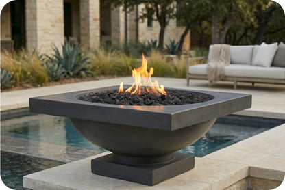 A square concrete fire pit table with a bowl-shaped base, filled with black lava rocks and lit flames, situated next to a swimming pool in an outdoor patio setting.