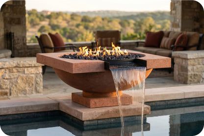 A freestanding round stone fire pit with black river rocks and visible flames, with a waterfall spout pouring water into a pool below. The fire pit sits on a patio beside a swimming pool, with outdoor seating and a softly blurred landscape in the background.