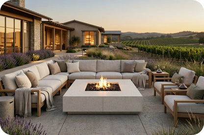 Outdoor patio furniture set arranged around a square fire pit table on a stone patio, with a modern house and vineyard visible in the background at sunset.