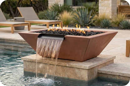 A rectangular outdoor fire pit with a copper-toned metal frame, black river rocks, and visible flames, designed with a waterfall feature that pours water into a pool. The fire pit is situated on a stone riser next to a swimming pool, with patio chairs and landscaping in the background.