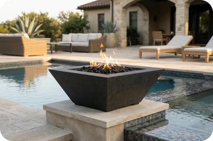 A modern square black fire pit with flames burning inside, set on a stone pedestal next to a swimming pool in an outdoor patio setting.