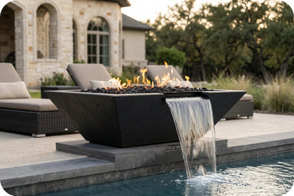 A modern black rectangular fire pit table with flowing flames and a waterfall spout into a pool, set on a patio in front of lounge chairs and a stone house at dusk.