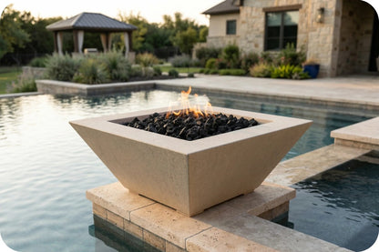 A square concrete fire pit table with a geometric design, filled with black fire rocks and a small flame, situated on a stone riser next to a swimming pool in a backyard setting.