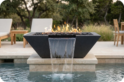 A modern black rectangular fire pit table with a waterfall design, set in a swimming pool with patio chairs and trees in the background.