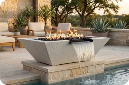 A modern concrete fire pit table with a waterfall feature, set on a stone patio beside a swimming pool with outdoor lounge chairs and plants in the background.