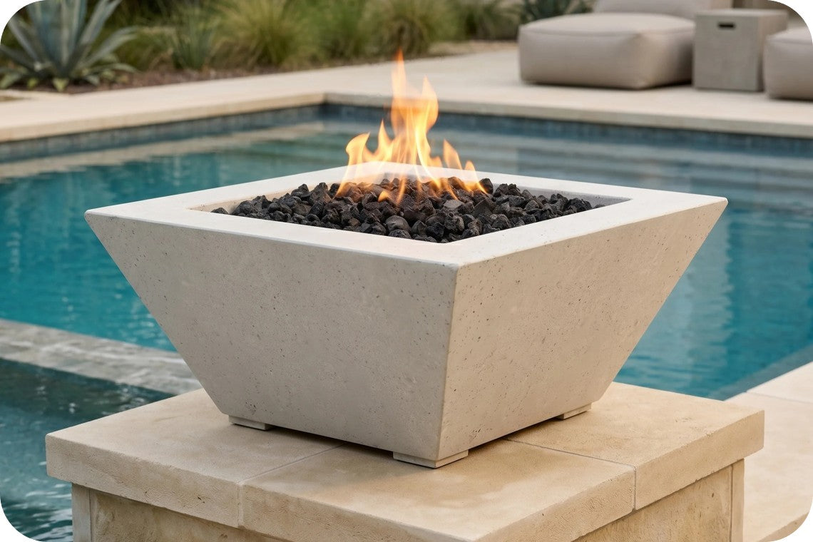 A square concrete fire pit table with a geometric design, filled with black lava rocks and active orange flames, situated outdoors on a stone pedestal next to a swimming pool.