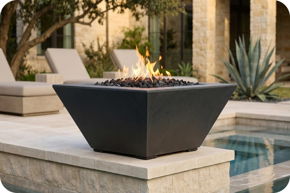 A modern black square fire pit table with a flame and black river rocks, set on a stone patio next to a swimming pool with a resort-style background.
