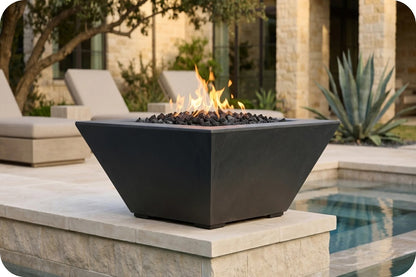 A modern black square fire pit table with a flame and black river rocks, set on a stone patio next to a swimming pool with a resort-style background.