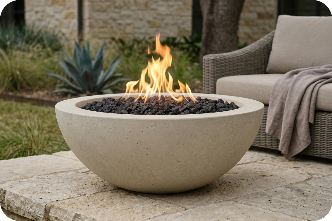 A round concrete fire pit bowl filled with black fire stones and active orange flames, set on a stone patio in an outdoor patio setting with a wicker sofa in the background.