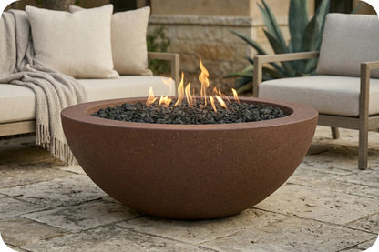 A round terracotta-colored outdoor fire pit bowl filled with black river rocks and lit flames, placed on a stone patio with patio furniture and plants in the background.