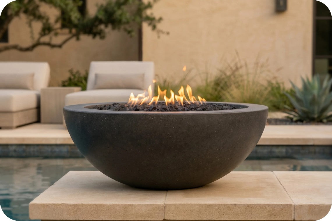 A dark charcoal-colored round outdoor fire pit bowl filled with black pebbles and active orange flames, set on a stone patio in front of a modern poolside patio background with lounge chairs.