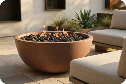 A large round terracotta fire pit table with black river rocks and visible orange flames, placed outdoors on a stone patio beside light beige patio furniture.