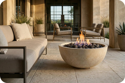A round concrete fire pit bowl with a glowing flame and dark stones, placed on a stone patio in a modern outdoor living room setting with a sofa and chairs.