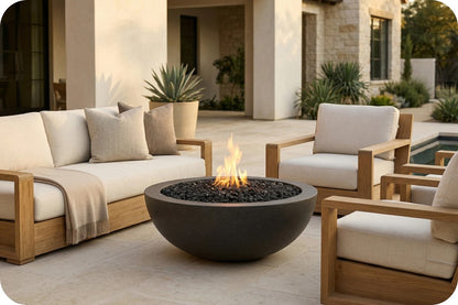 Outdoor patio furniture set with a round dark fire pit bowl in the center, flanked by two light wood armchairs and a matching sofa with cream cushions, in front of a modern house.