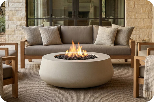 A round concrete fire pit table with a glowing fire in the center, placed in front of a modern outdoor sofa with neutral cushions on a woven rug.