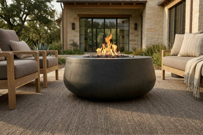 A round dark gray concrete fire pit with a glowing fire, situated in the center of an outdoor patio with wooden seating and a stone house in the background.