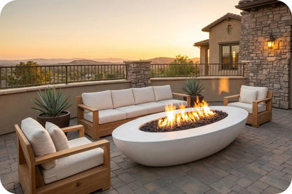 Outdoor patio furniture set with a white oval fire pit table, a wooden three-seat sofa, and two matching armchairs with cream cushions, arranged on a stone patio at sunset with a house and hills in the background.
