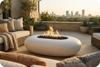 A modern oval outdoor fire pit table with a white stone-like finish, set on a patio with a city skyline in the background. The fire burns brightly with orange and yellow flames, surrounded by neutral-toned patio furniture and potted plants.