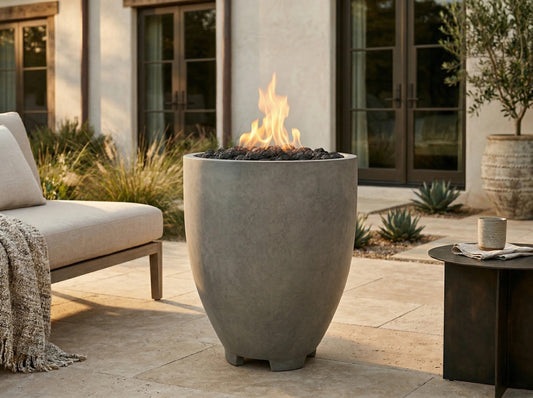 A large concrete outdoor fire pit with a glowing flame and dark stones, placed on a patio beside a light beige lounge chair and a small dark side table.