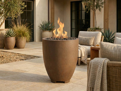 A tall, matte brown outdoor fire pit with a glowing flame, set on a stone patio in a modern patio lounge setting.
