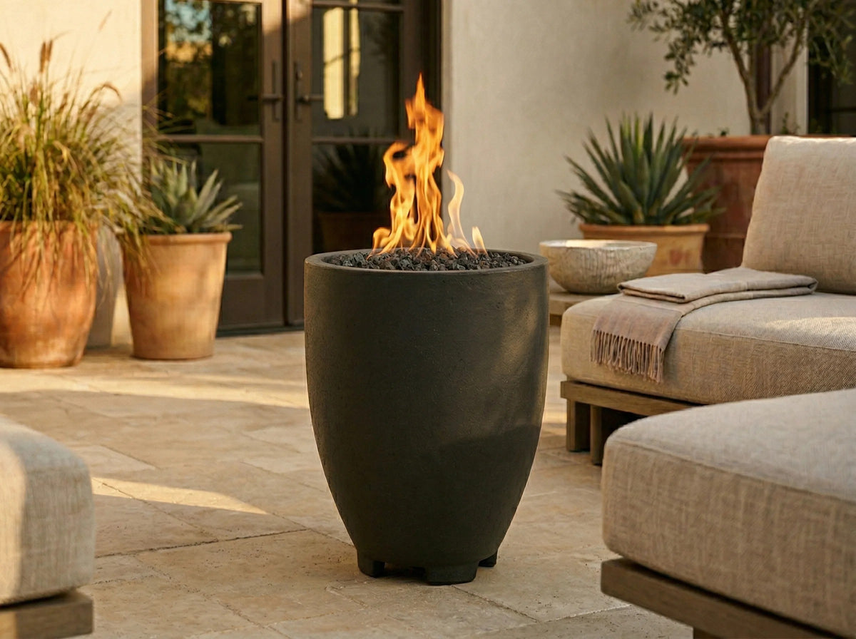 A modern outdoor fire pit with a matte black conical pot and active orange flames, set on a stone patio next to beige outdoor seating.