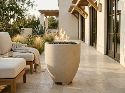 A large beige concrete outdoor fire pit with visible flames and dark stones inside, placed on a stone patio beside a lounge chair in a modern patio setting.