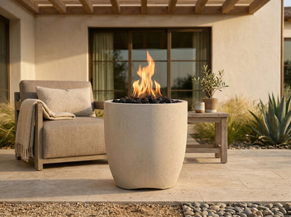 A modern outdoor patio scene featuring a tall, cylindrical concrete fire pit with a glowing orange flame and black stones inside, positioned in front of a beige lounge chair and a small wooden side table.