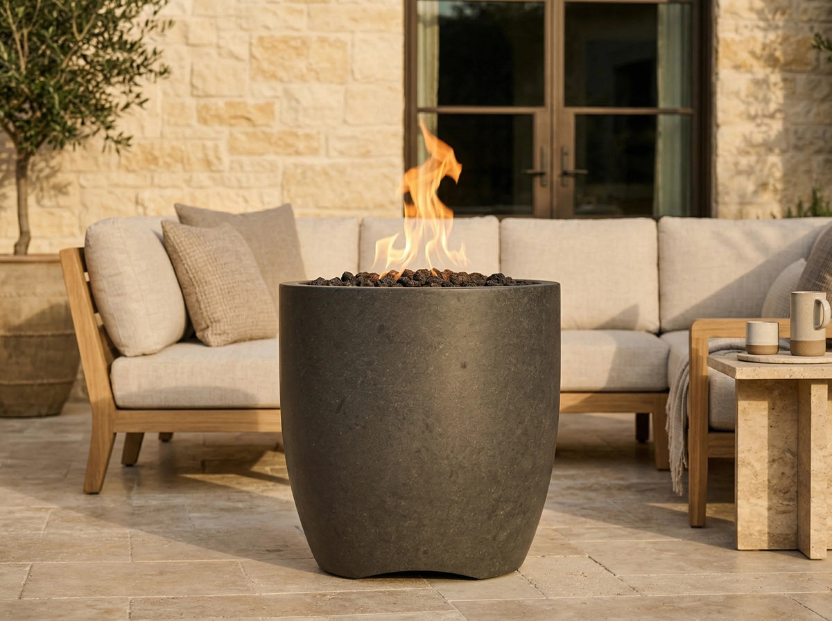 A dark charcoal gray concrete fire pit with a live flame burning inside, set on a stone patio in front of a light beige outdoor sofa.