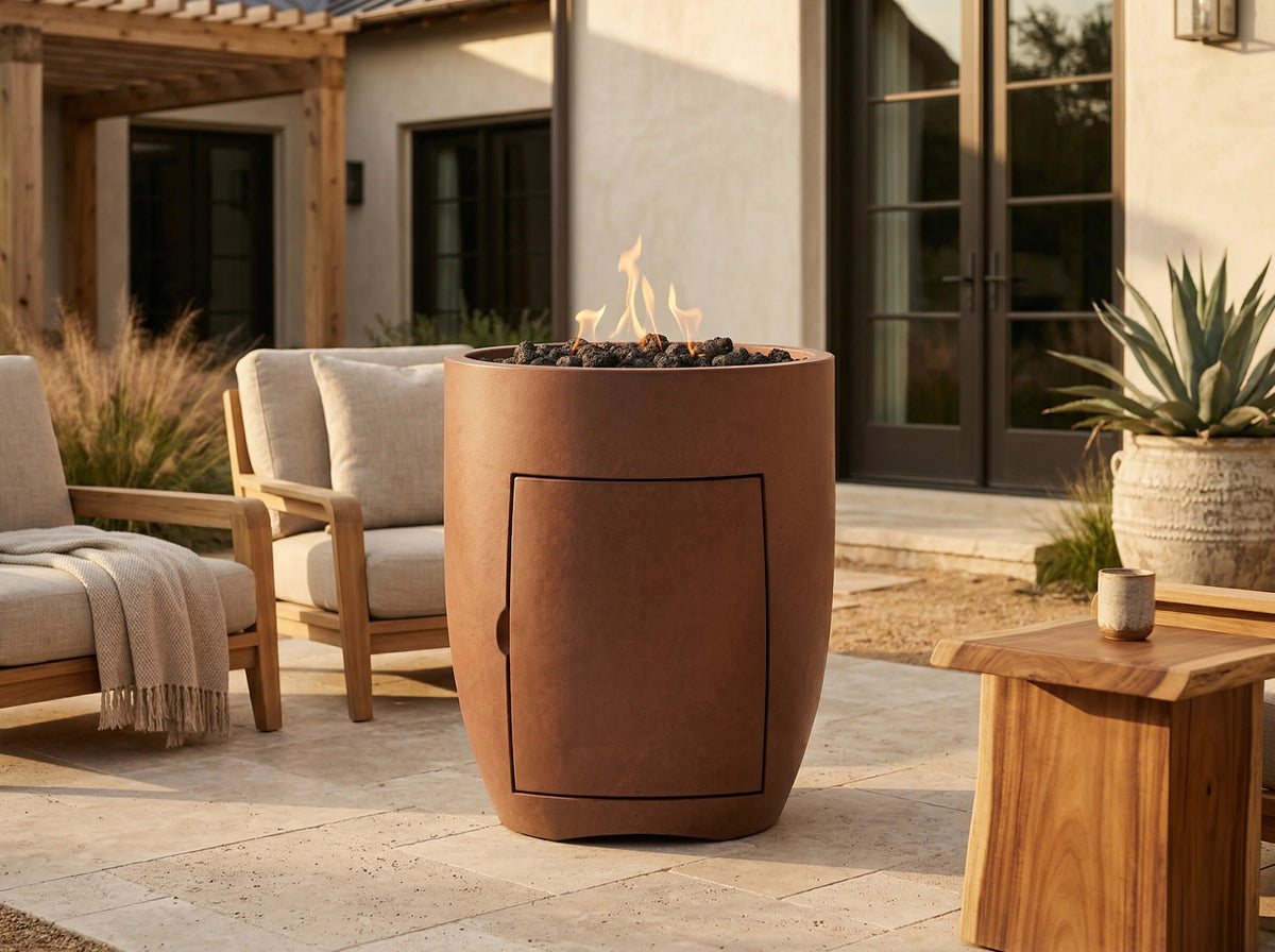 A tall cylindrical outdoor fire pit table with a warm terracotta finish, filled with dark river rocks and small flames, set on a patio next to lounge chairs and a wooden side table.