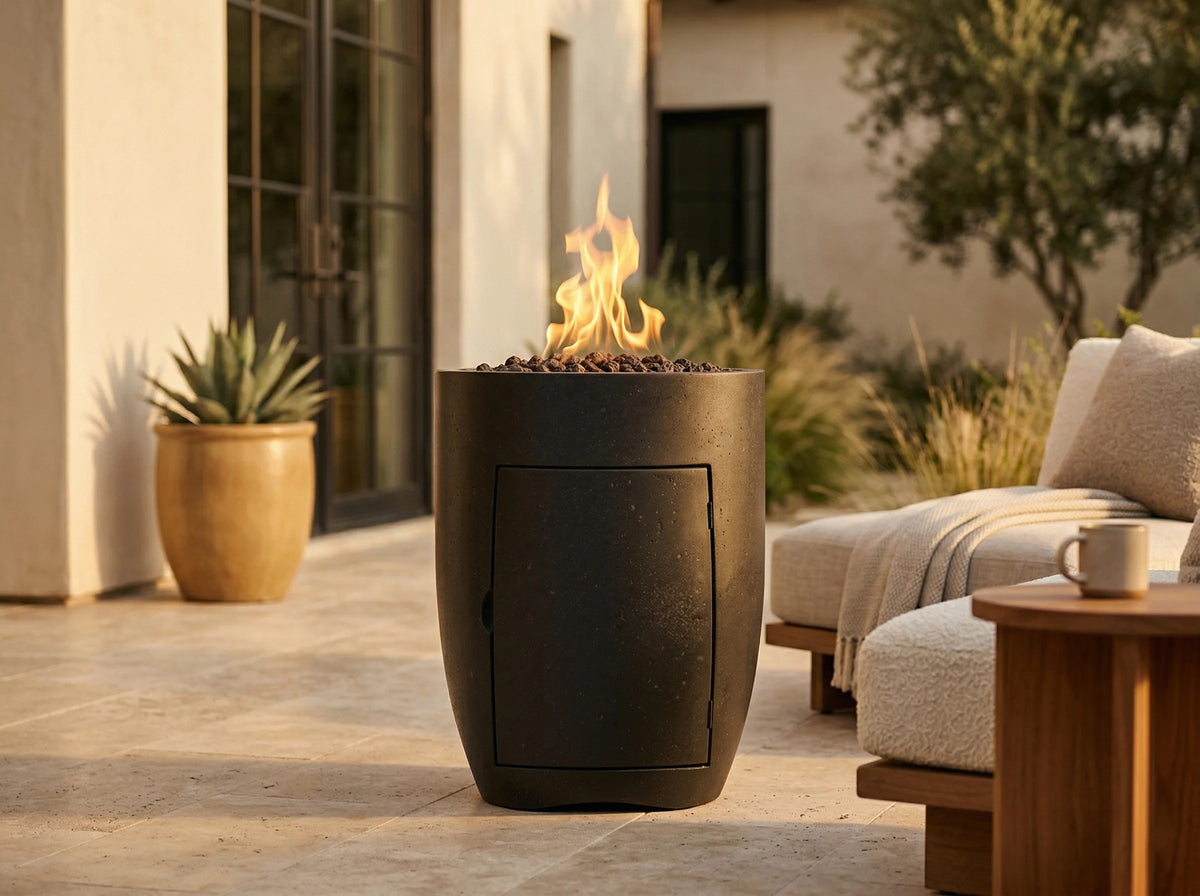 A modern black outdoor fire pit table with a glowing flame, situated on a stone patio next to a beige lounge chair and a wooden side table.