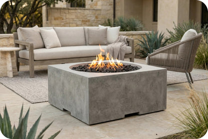 A square concrete fire pit table with a burning fire, set on a stone patio in an outdoor patio lifestyle scene with a beige sofa and a wicker armchair.