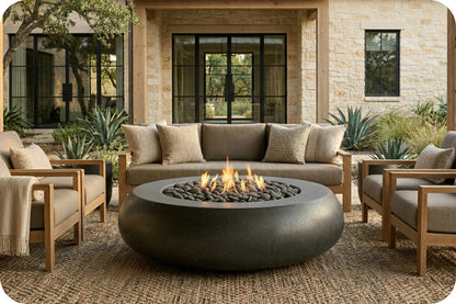Outdoor patio furniture set featuring a round dark gray fire pit table with a lit flame, a beige sofa, two matching armchairs, and a woven area rug in front of a stone house.