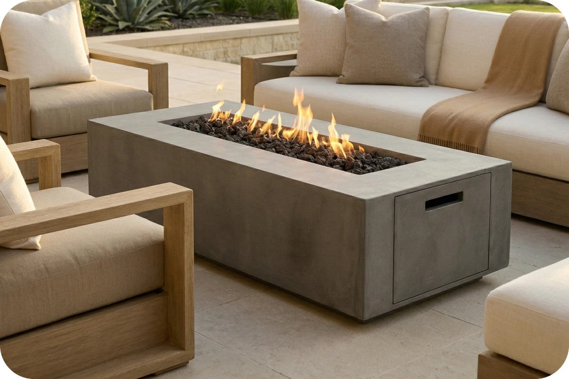 A rectangular concrete outdoor fire pit table with a visible flame and dark river rocks, placed in a patio seating area with cream sofas and wooden chairs.