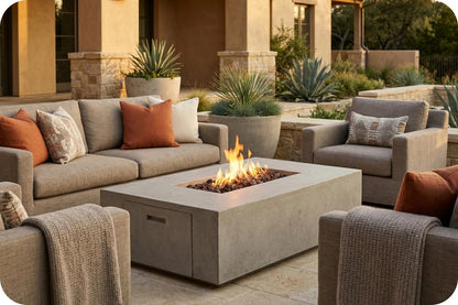 Outdoor patio furniture set with a rectangular concrete fire pit table in the center, surrounded by matching gray sectional sofas and armchairs with orange and beige throw pillows, set in front of a warm-toned stucco building with potted plants.