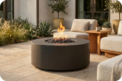 A round concrete fire pit table with a visible flame and black stones, placed on a patio in an outdoor living room setting with lounge chairs and plants.