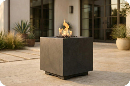 A modern square outdoor fire pit with a matte dark charcoal finish, featuring active orange and yellow flames rising from black stones, set on a stone patio in front of a house.