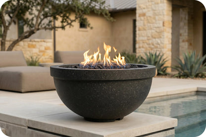 A round black stone fire pit bowl with active orange and yellow flames, set on a light stone patio next to a swimming pool with a modern house and outdoor seating in the background.