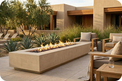 A modern rectangular concrete outdoor fire pit with active flames, situated on a stone patio between contemporary beige lounge chairs and desert landscaping.